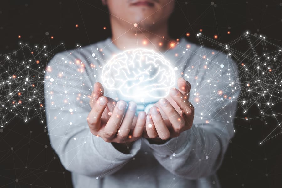 Manos sostienen un cerebro humano luminoso ilustrado con red de puntos y conexiones sobre fondo oscuro, símbolo de la rehabilitación logopédica en enfermedades neurológicas.
