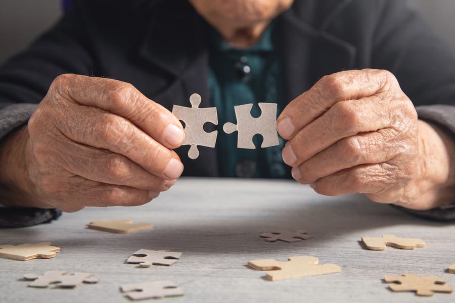 Manos arrugadas de persona mayor unen dos piezas de puzzle de cartón sobre la mesa con piezas dispersas, ejercicio de estimulación cognitiva en Alzheimer y demencias.