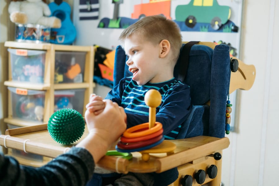 Niño sonriente sentado en silla adaptada de madera juega con pelota sensorial verde y aros apilables sobre bandeja, sesión de estimulación temprana para parálisis cerebral infantil.